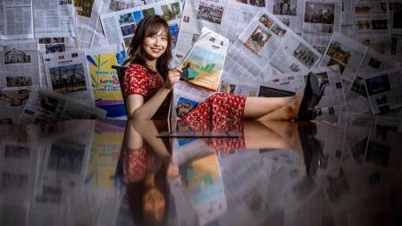 Heesoo Jang reading a newspaper in front of background filled up with newspaper clippings.